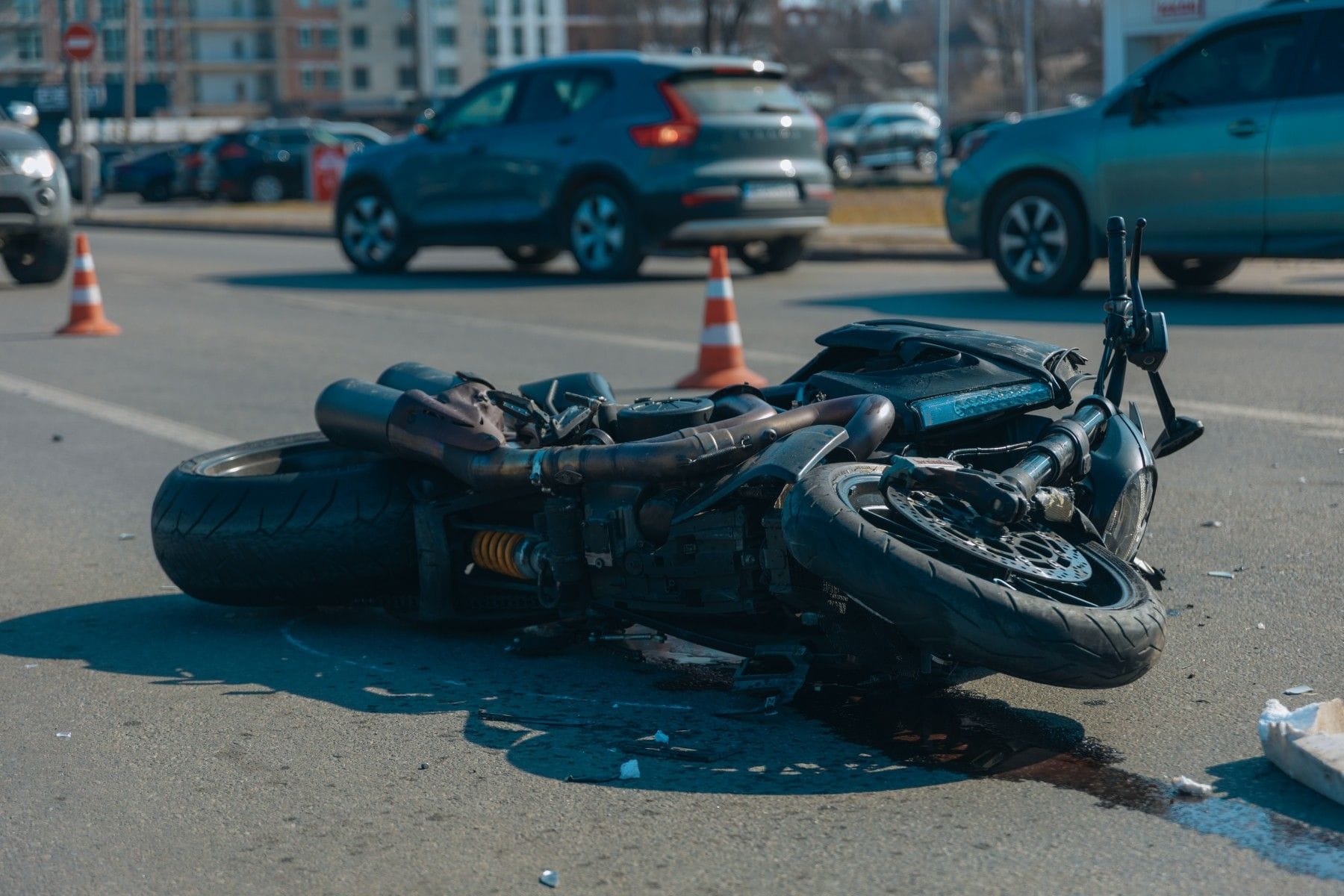 A motorcycle lies on its side in a roadway near traffic cones, with cars in the background.