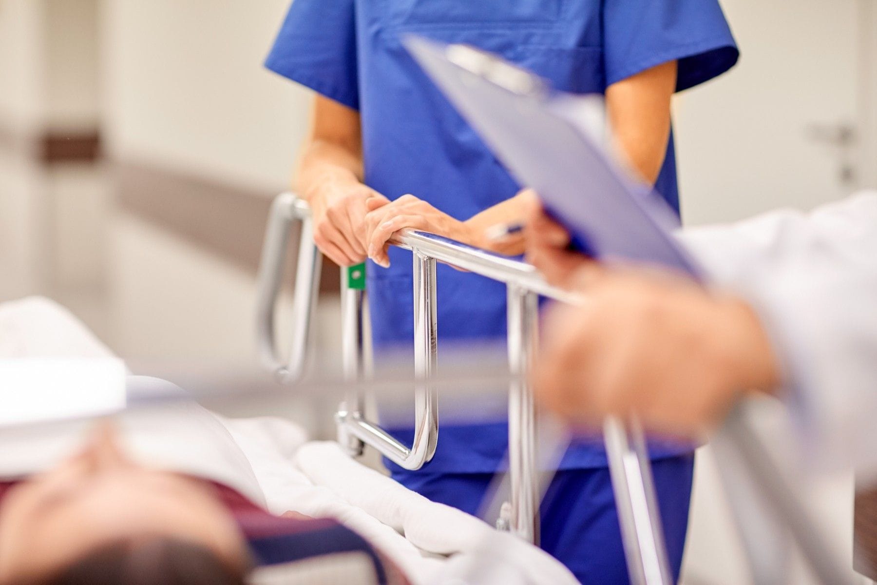 A clinician in blue scrubs stands beside a patient in a hospital bed, holding the patient’s hand near a metal bedside rail.