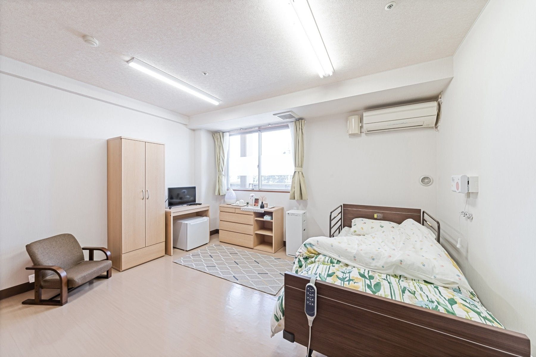 An empty nursing home room with an adjustable bed, armchair, wardrobe, television, and window.