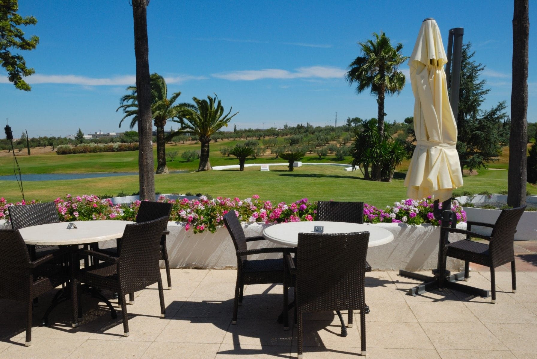 Outdoor restaurant patio beside a landscaped golf course, with tables, chairs, flowers, palm trees, and a closed umbrella.